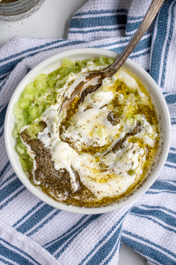 Small white bowl over a blue towel with a spoon mixing together the ingredients for homemade tzatziki sauce showing swirls of olive oil and ill in yogurt and cucumbers.