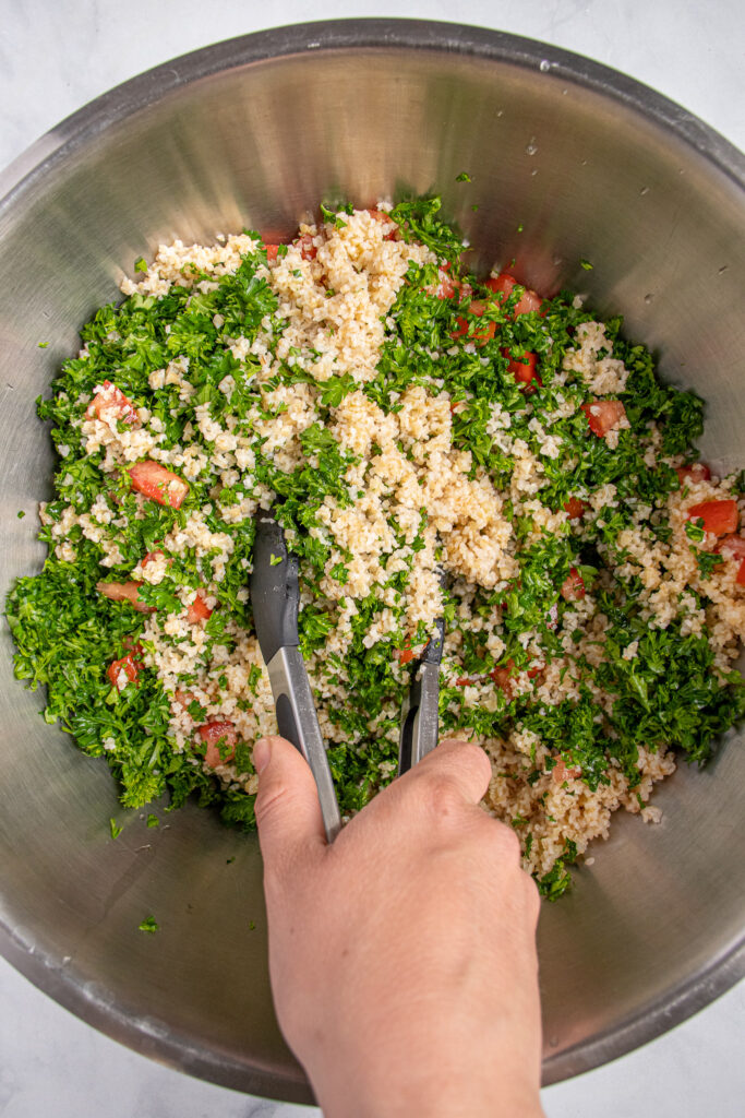 A person using tongs to mix the ingredients of tabbouleh salad together including parsley, bulgur, and tomatoes.