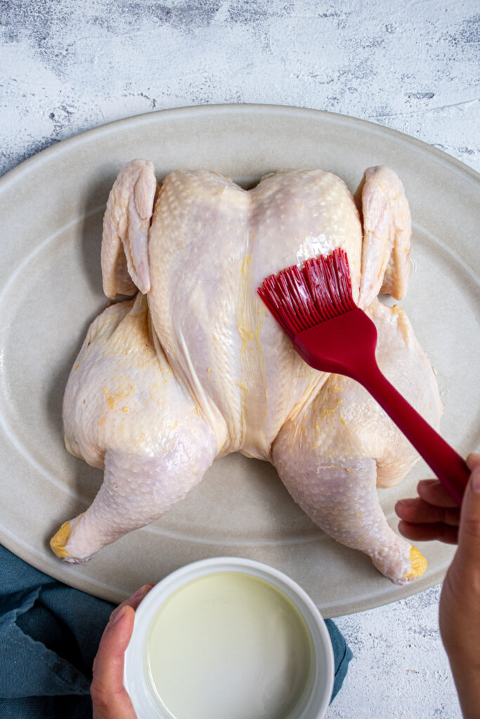 A raw spatchcocked chicken on a platter being oiled with a silicone brush.