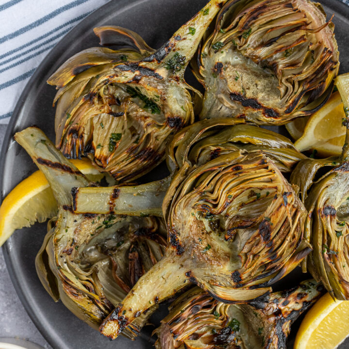 A dark platter full of stacked grilled artichoke halves with lemon wedges.