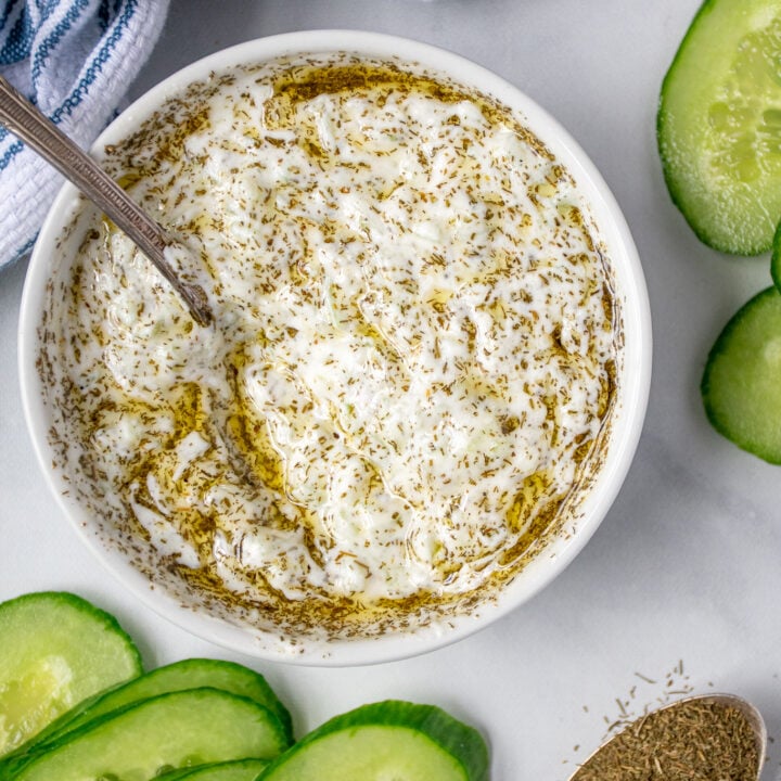 Overhead view of a small white bowl next to some cucumbers and a spoon, in the bowl is homemade tzatziki sauce.