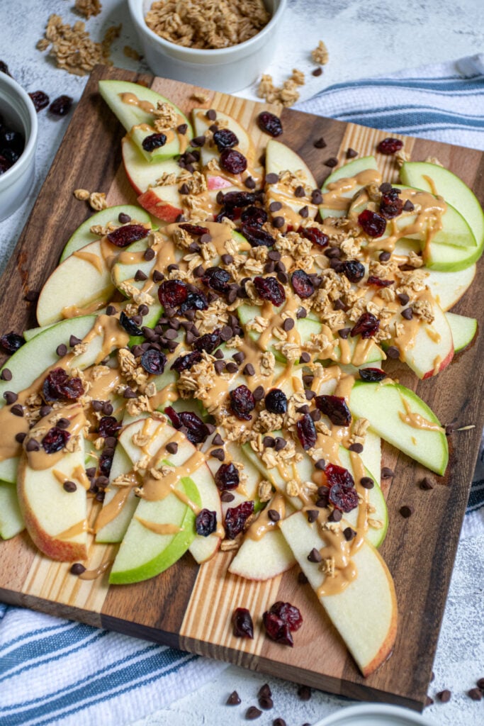 Cutting board loaded with apple slices piled high with peanut butter, granola, dried cranberries, and chocolate chips.