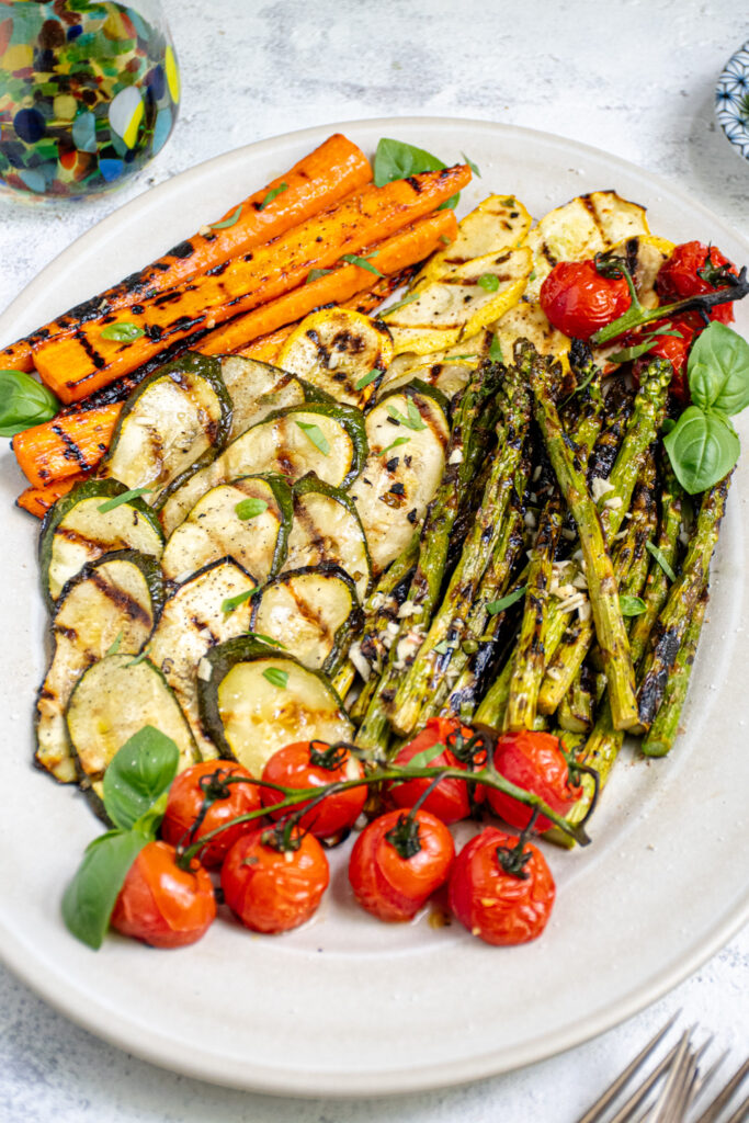 Angled view of a white platter filled with marinated and grilled vegetables including zucchini, asparagus, and tomatoes.