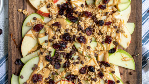 Overhead view of a wood cutting board piled high with apple slices covered in peanut butter, granola, dried cranberries, and mini chocolate chips.