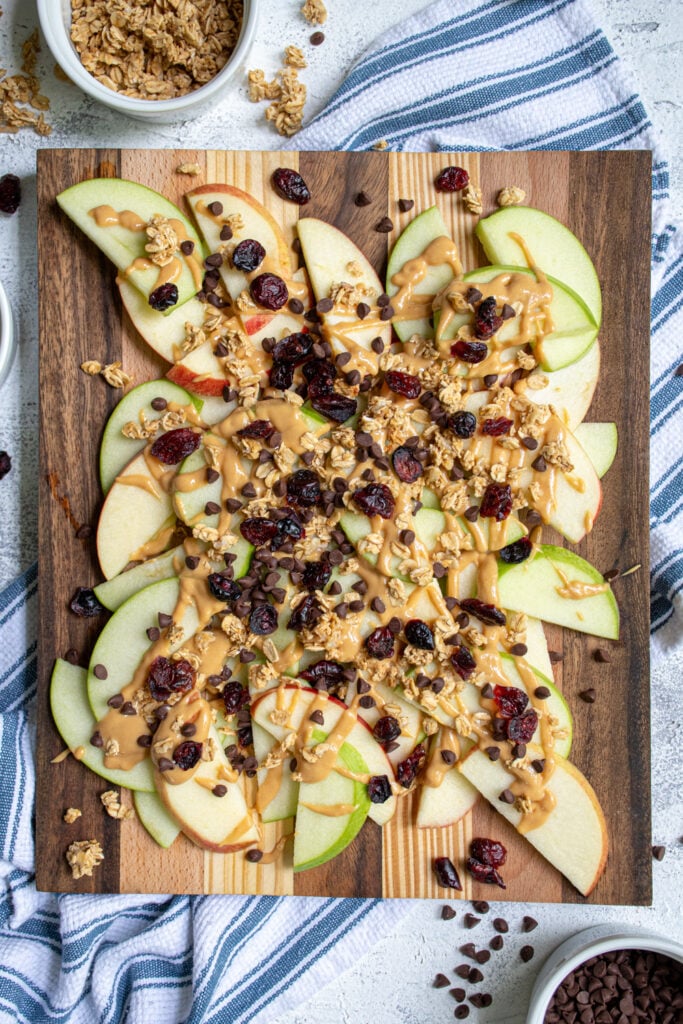 Overhead view of a wood cutting board piled high with apple slices covered in peanut butter, granola, dried cranberries, and mini chocolate chips. 