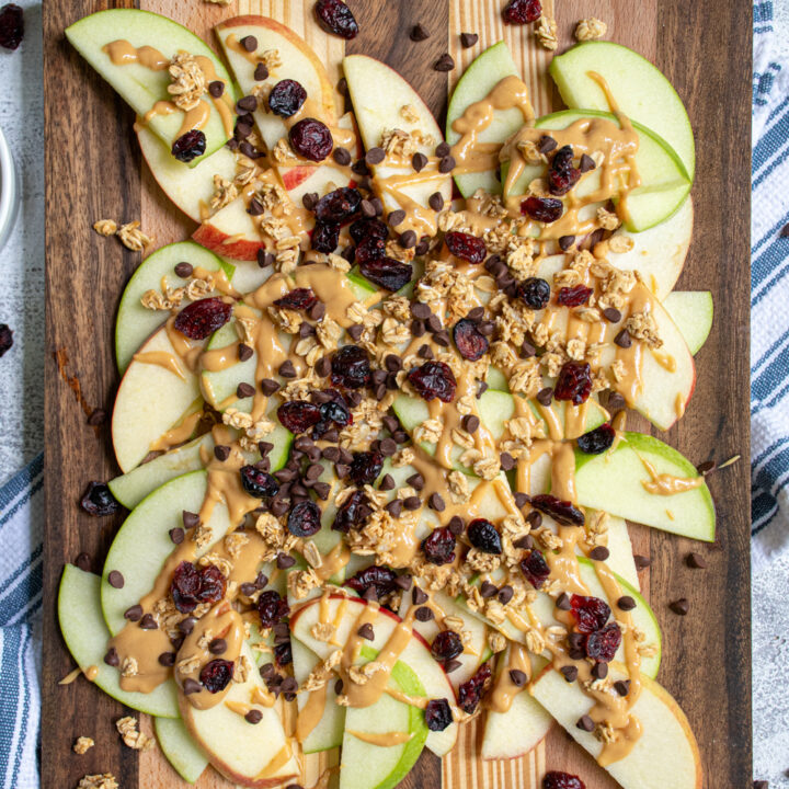 Overhead view of a wood cutting board piled high with apple slices covered in peanut butter, granola, dried cranberries, and mini chocolate chips.