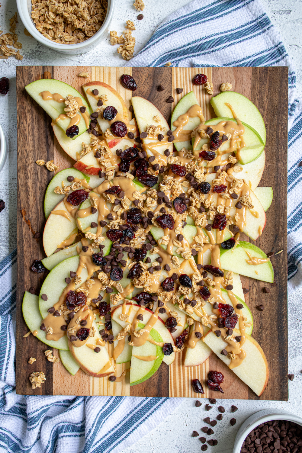 Overhead view of a wood cutting board piled high with apple slices covered in peanut butter, granola, dried cranberries, and mini chocolate chips.