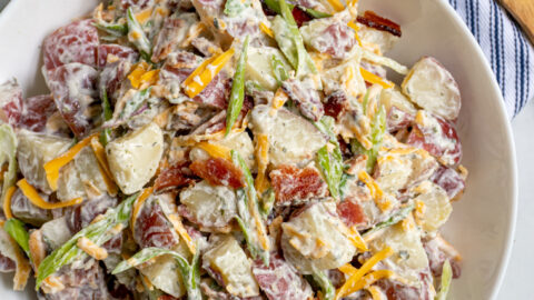 A large white bowl filled with healthier bacon ranch potato salad filled with bacon bits, cheddar cheese, and sliced green onions.