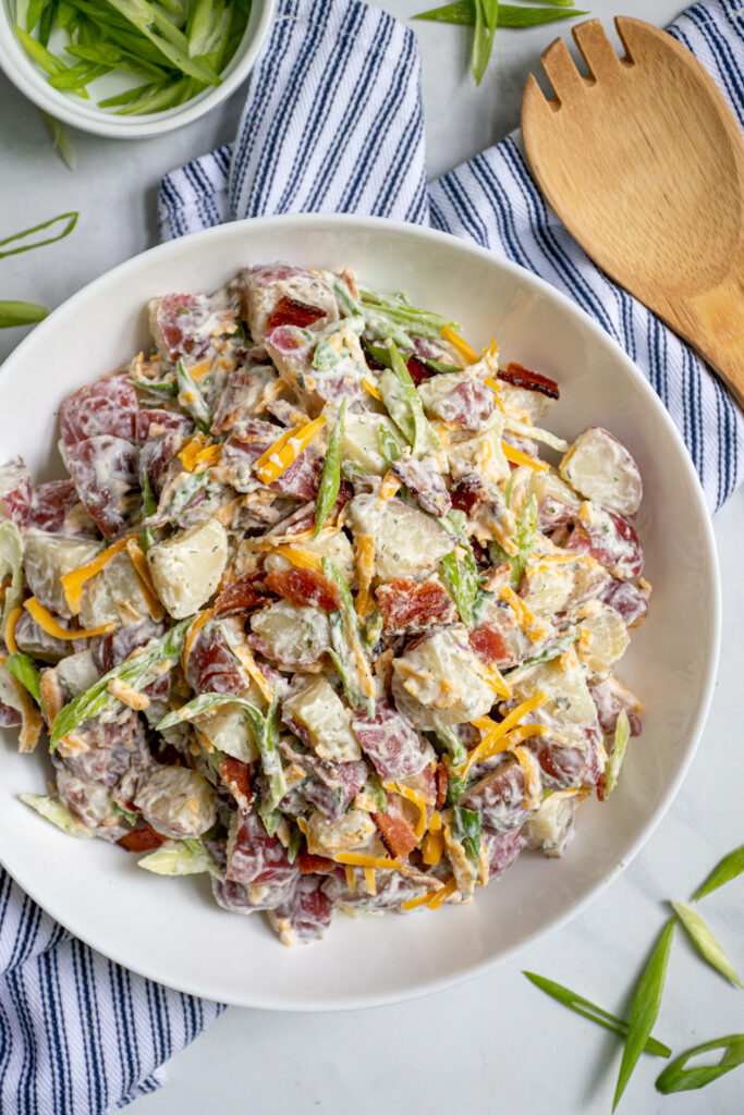 A large white bowl filled with healthier bacon ranch potato salad filled with bacon bits, cheddar cheese, and sliced green onions.