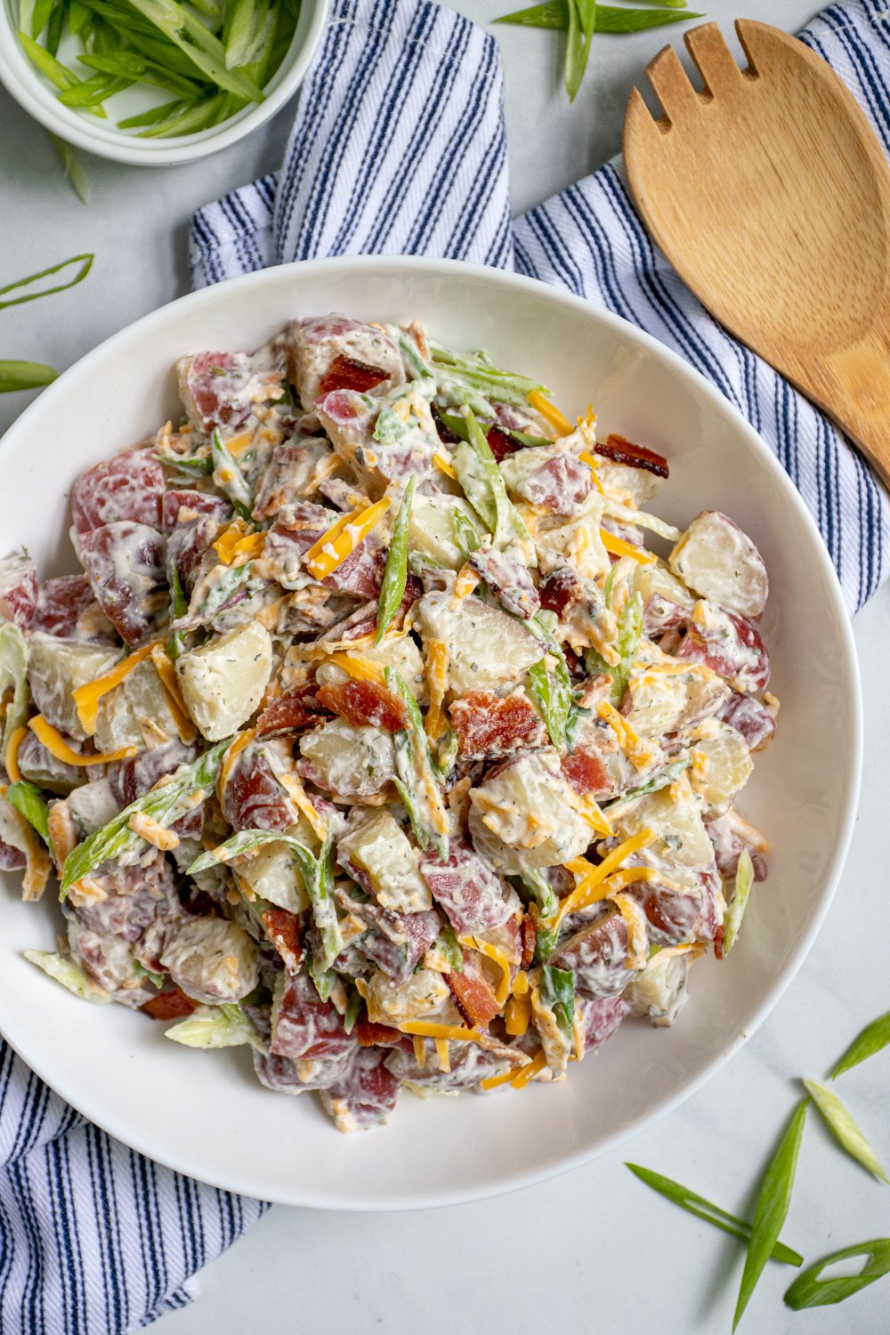 A large white bowl filled with healthier bacon ranch potato salad filled with bacon bits, cheddar cheese, and sliced green onions.