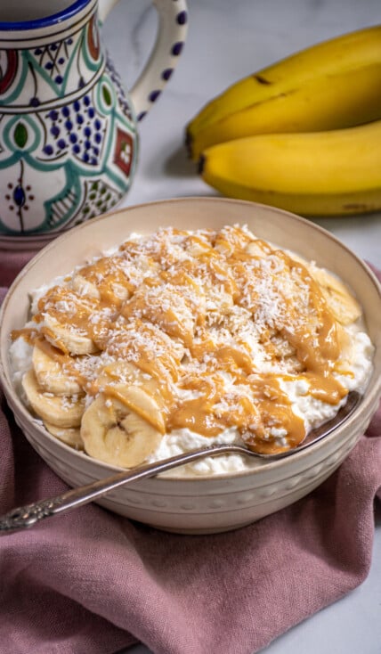 White bowl gilled with cottage cheese and topped with banana slices, peanut butter, and shredded coconut.