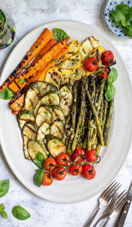 Overhead view of a white platter filled with marinated grilled veggies including zucchini, asparagus, carrots, and tomatoes.