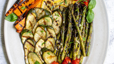 Overhead view of a white platter filled with marinated grilled veggies including zucchini, asparagus, carrots, and tomatoes.