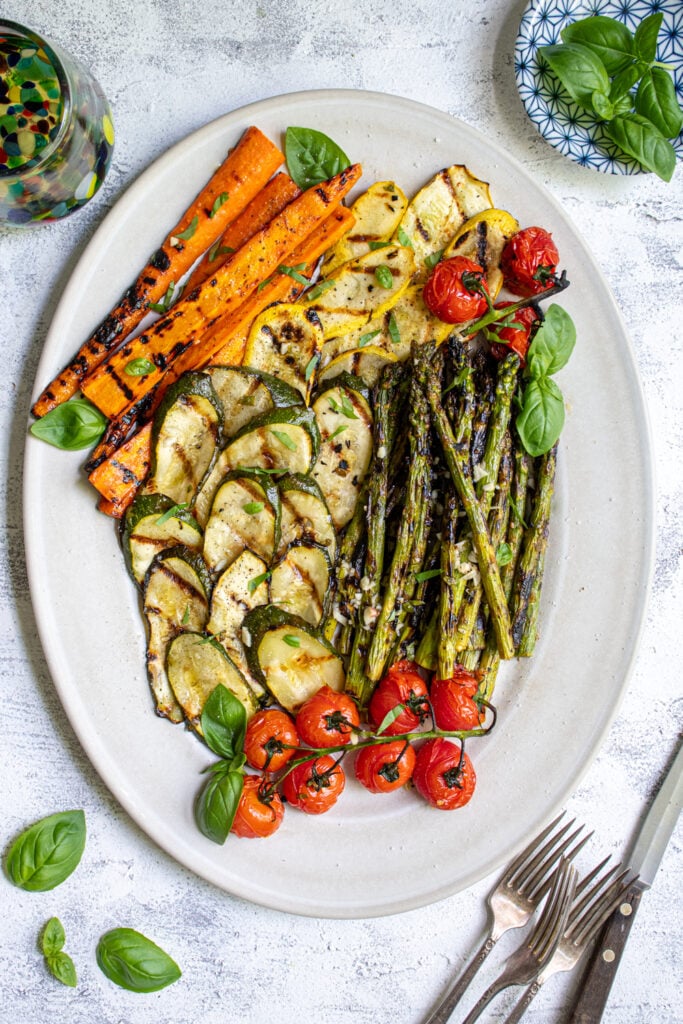 Overhead view of a white platter filled with marinated grilled veggies including zucchini, asparagus, carrots, and tomatoes.