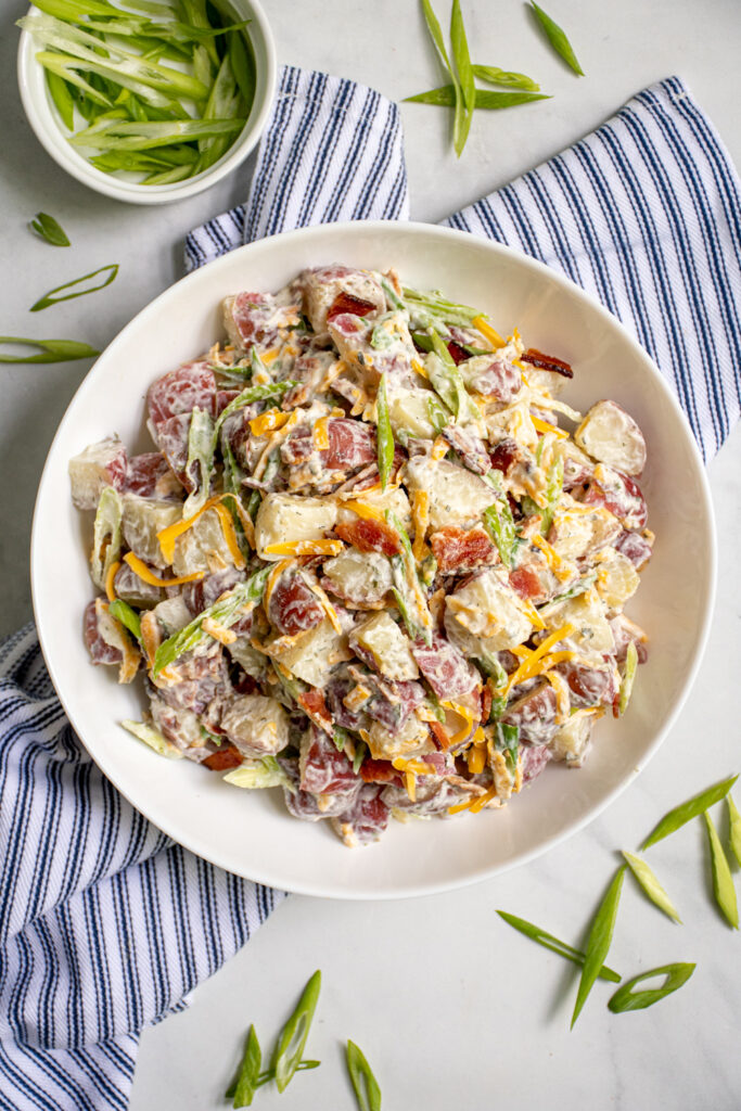 View from overhead of a large white bowl filled with bacon ranch potato salad the bowl is sitting on a blue and white towel and is surrounded by sliced green onions.