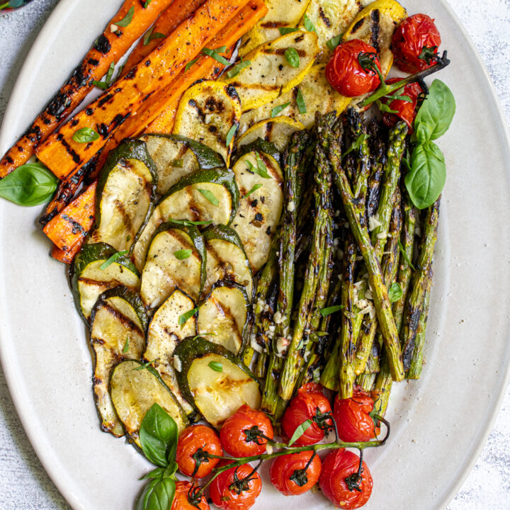 Marinated and grilled zucchini, asparagus, carrots, and tomatoes on a platter covered in fresh basil leaves.