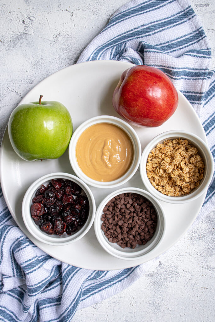 Ingredients for apple nachos sitting on a plate including apples, peanut butter, granola, dried cranberries, and mini chocolate chips.