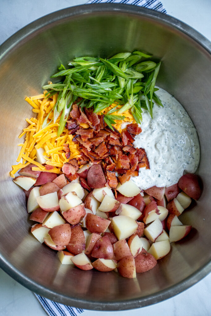 All the ingredients in a large stainless steel serving bowl ready to be tossed to gather to make bacon ranch potato salad.