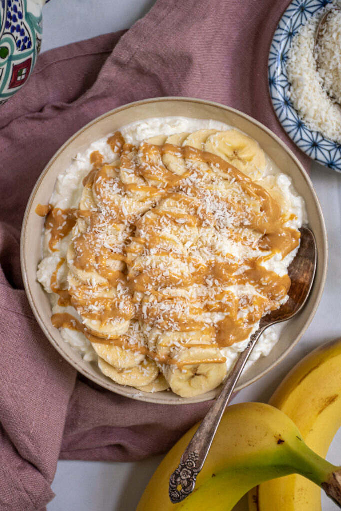 Close up of a bowl filled with cottage cheese and topped with banana slices and drizzled peanut butter with shredded coconut flakes.