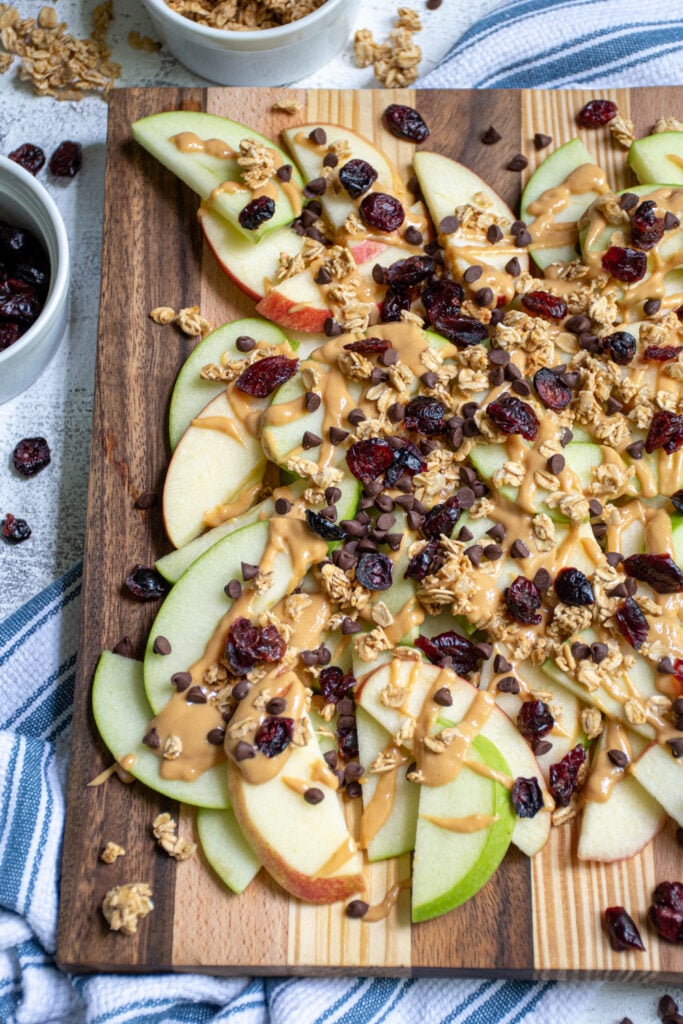 Wood cutting board layered with apple slices, peanut butter, granola, dried cranberries, and mini chocolate chips.