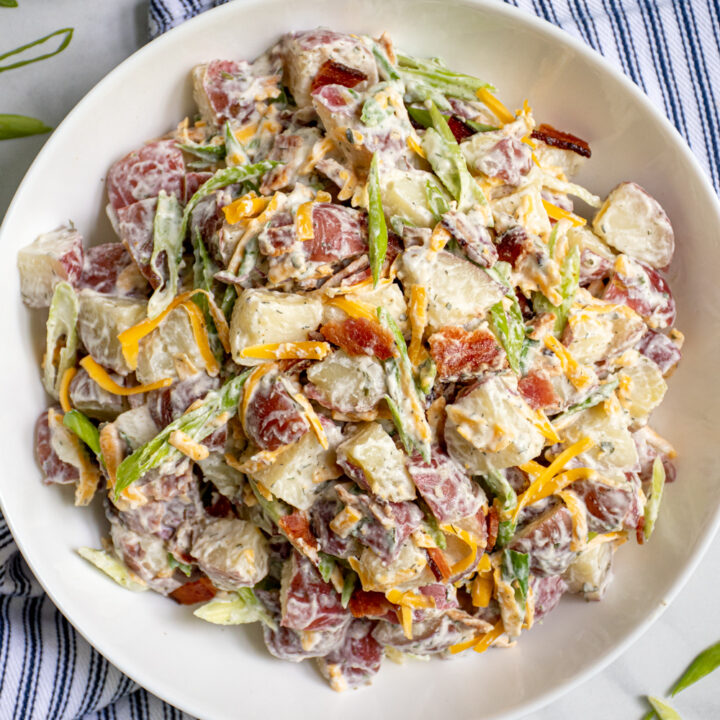 Close up of bacon ranch potato salad in a large white bowl that is sitting on a blue and white stripped towel.