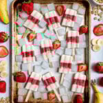 Sheet tray full of red and white parfait popsicles and ice cubes surrounded bu sliced strawberries, bananas, and bits of granola.