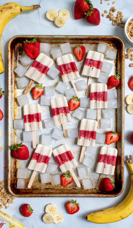 Sheet tray full of red and white parfait popsicles and ice cubes surrounded bu sliced strawberries, bananas, and bits of granola.