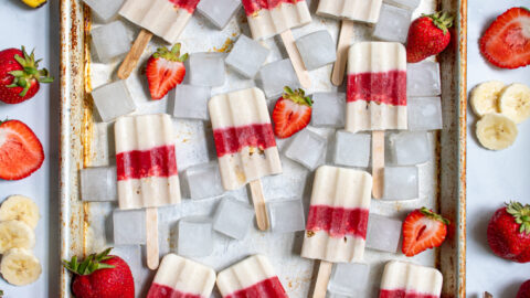Sheet tray full of red and white parfait popsicles and ice cubes surrounded bu sliced strawberries, bananas, and bits of granola.