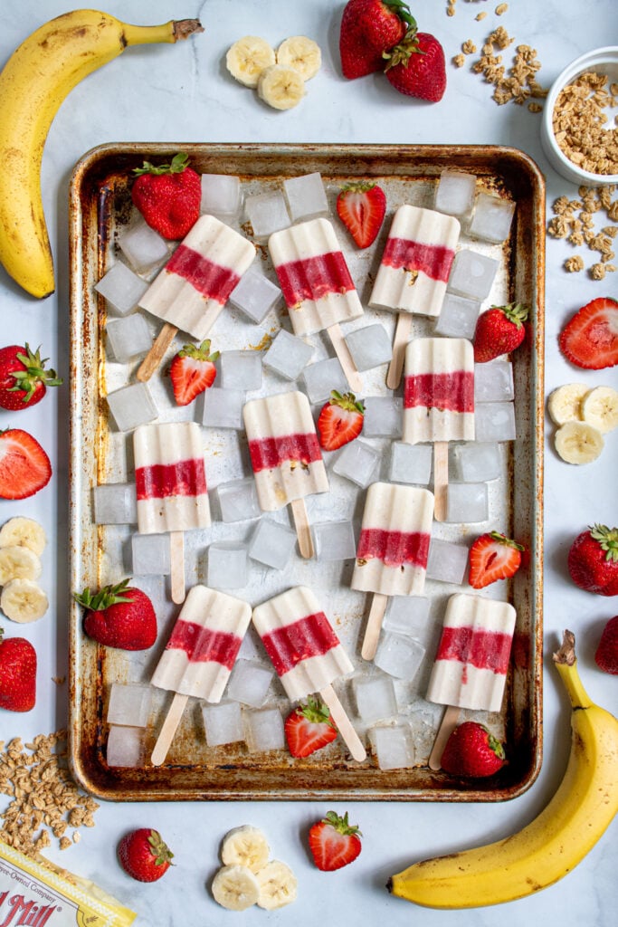 Sheet tray full of red and white parfait popsicles and ice cubes surrounded bu sliced strawberries, bananas, and bits of granola.
