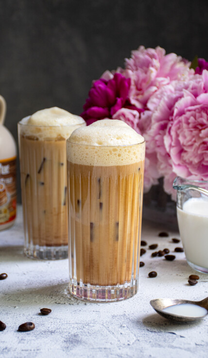 A glass filled with ice and salted maple cold brew coffee sitting on a table top next to another drink and a vase of pink flowers.