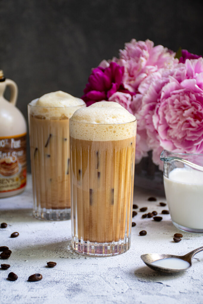 A glass filled with ice and salted maple cold brew coffee sitting on a table top next to another drink and a vase of pink flowers.