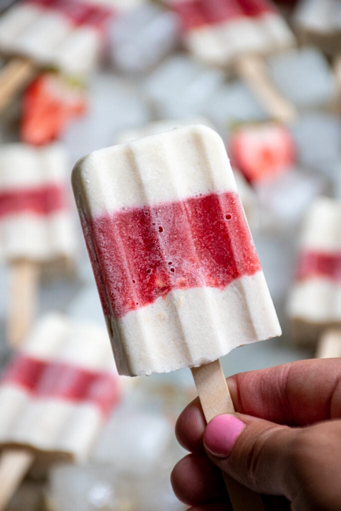 Hand holding up a popsicle stick of a parfait popsicle showing white and red layers.