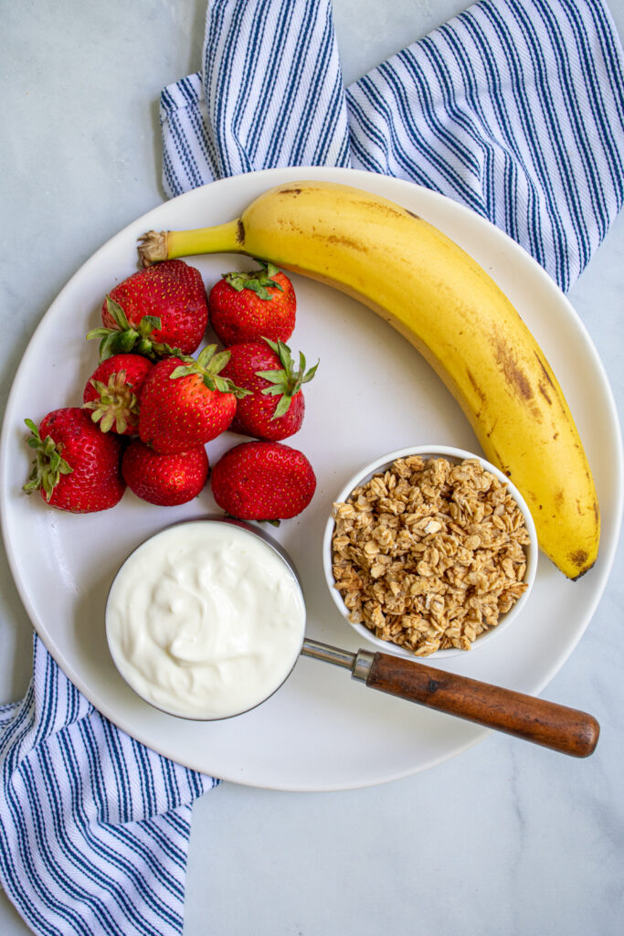 Ingredients to make parfait popsicles on a white plate including strawberries, a banana, yogurt, and granola.