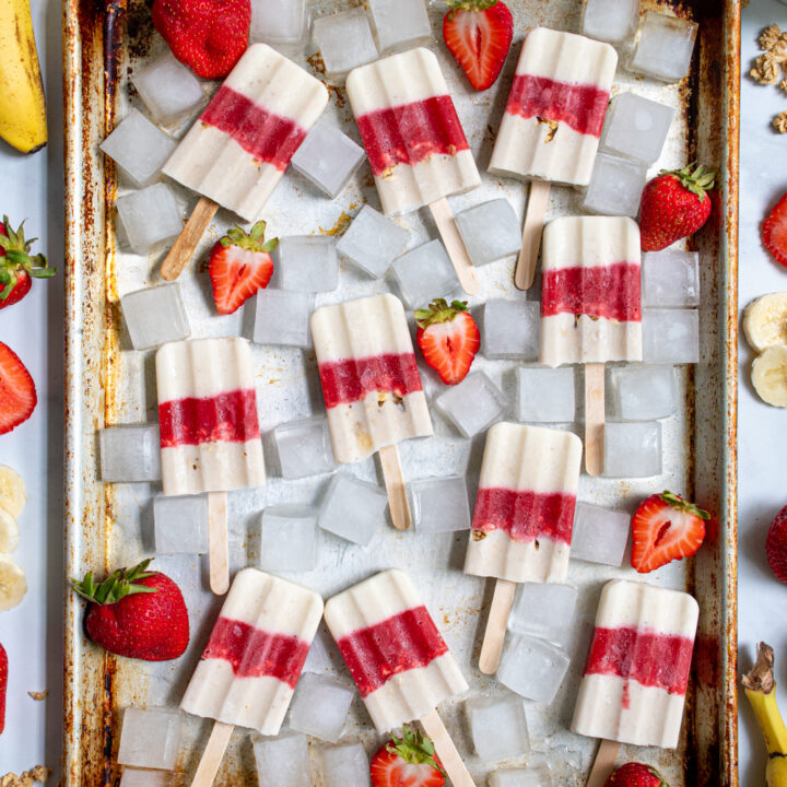 Overhead view of a sheet pan filled with red and white parfait breakfast popsicles surrounded by ice cubes and sliced strawberries.