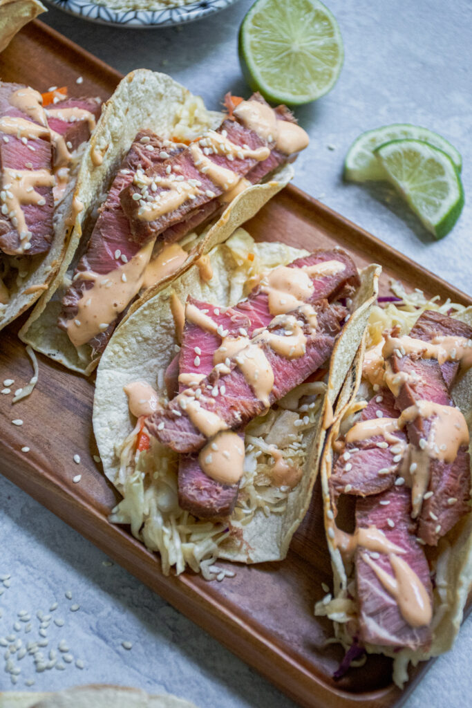 Angled view of the tuna steak tacos showing off the sliced tuna steak overtop a bed of coleslaw and topped with sriracha mayo and sesame seeds.