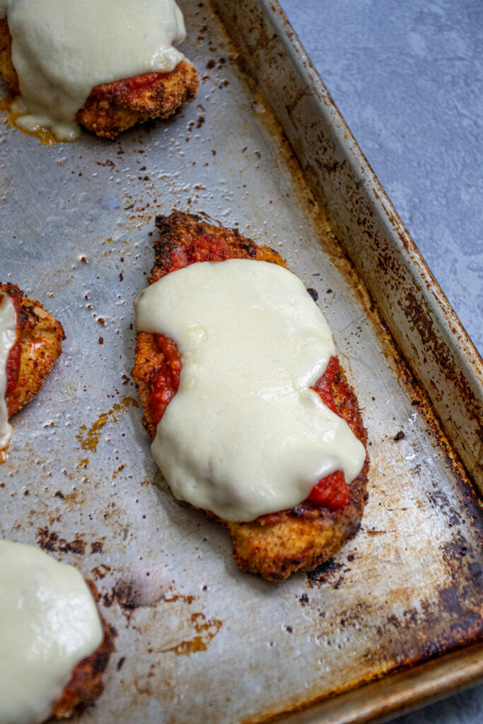 A close of a baked chicken parm on a sheet tray with marinara sauce and mozzarella cheese.