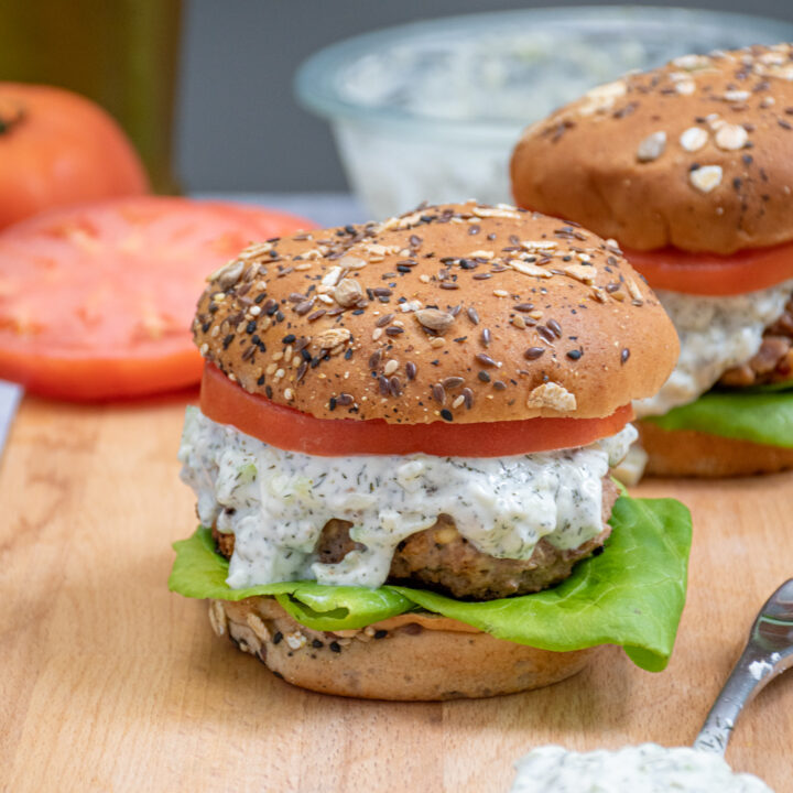 A greek turkey burger topped with tzatziki sauce, lettuce, and tomato sitting on a cutting board black next to a spoon of tzatziki.