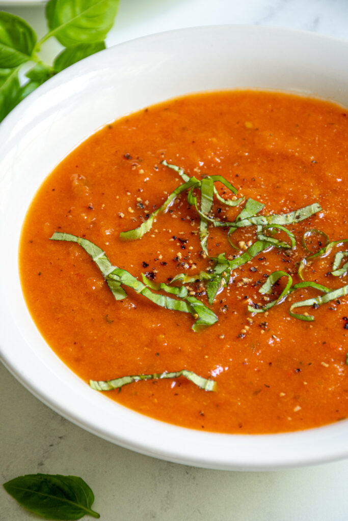 Close up of a white bowl of red-orange roasted red pepper and tomato soup garnished with cracked black pepper and fresh basil.