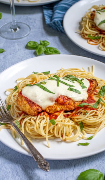 A white plate piled high with spaghetti topped with an Oven Baked Chicken Parmesan topped with melted mozzarella and garnished with pieces of fresh basil.