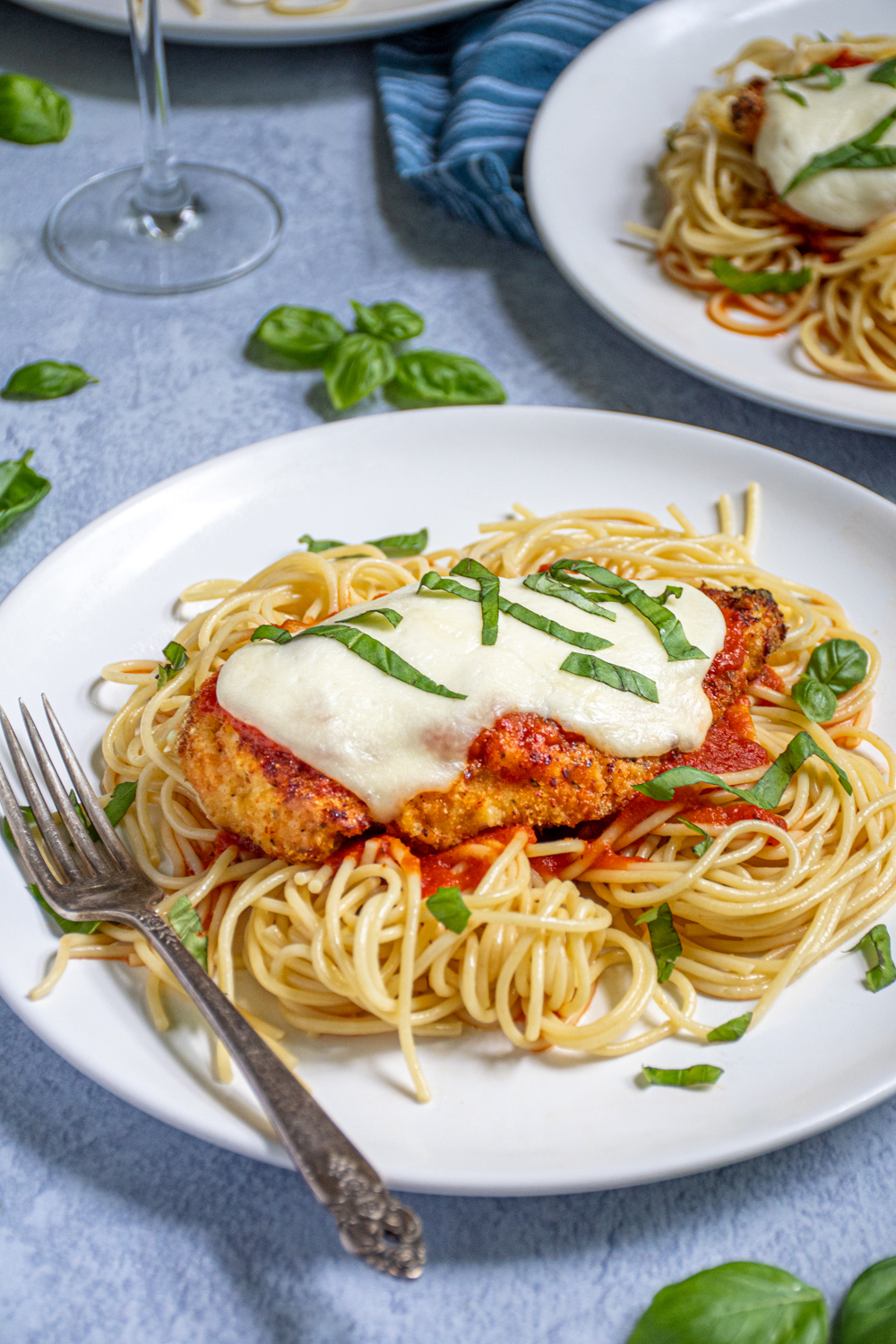 A white plate piled high with spaghetti topped with an Oven Baked Chicken Parmesan topped with melted mozzarella and garnished with pieces of fresh basil.