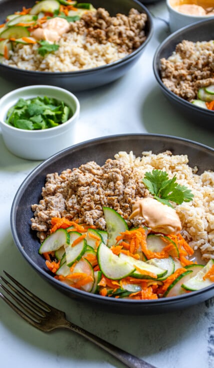 A black bowl filled with brown rice, ground pork, and pickled vegetables making a Banh Mi bowl garnished with sriracha mayo and cilantro.