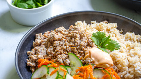 A black bowl filled with brown rice, ground pork, and pickled vegetables making a Banh Mi bowl garnished with sriracha mayo and cilantro.