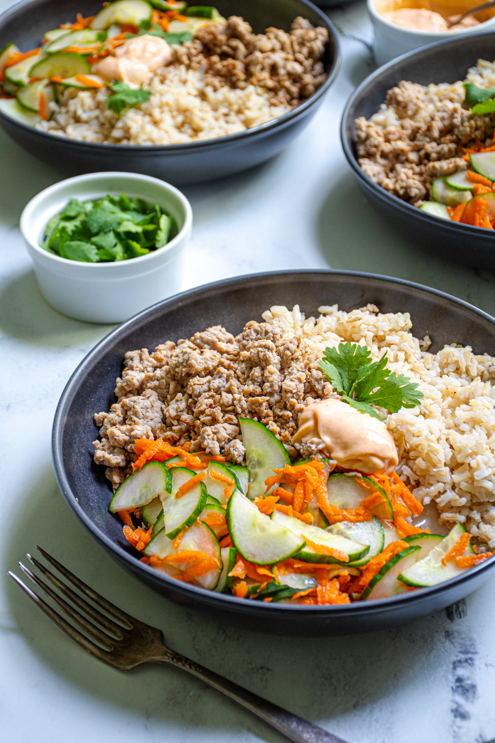 A black bowl filled with brown rice, ground pork, and pickled vegetables making a Banh Mi bowl garnished with sriracha mayo and cilantro.