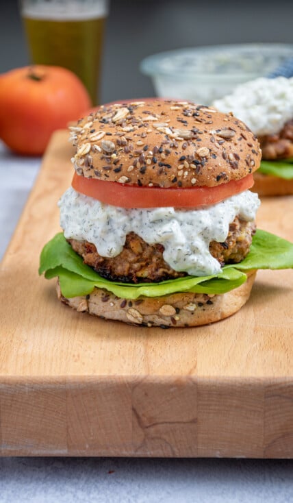 A Mediterranean Turkey Burger topped with Tzatziki sauce, tomatoes, and lettuce sitting on a butcher block next to another turkey burger.