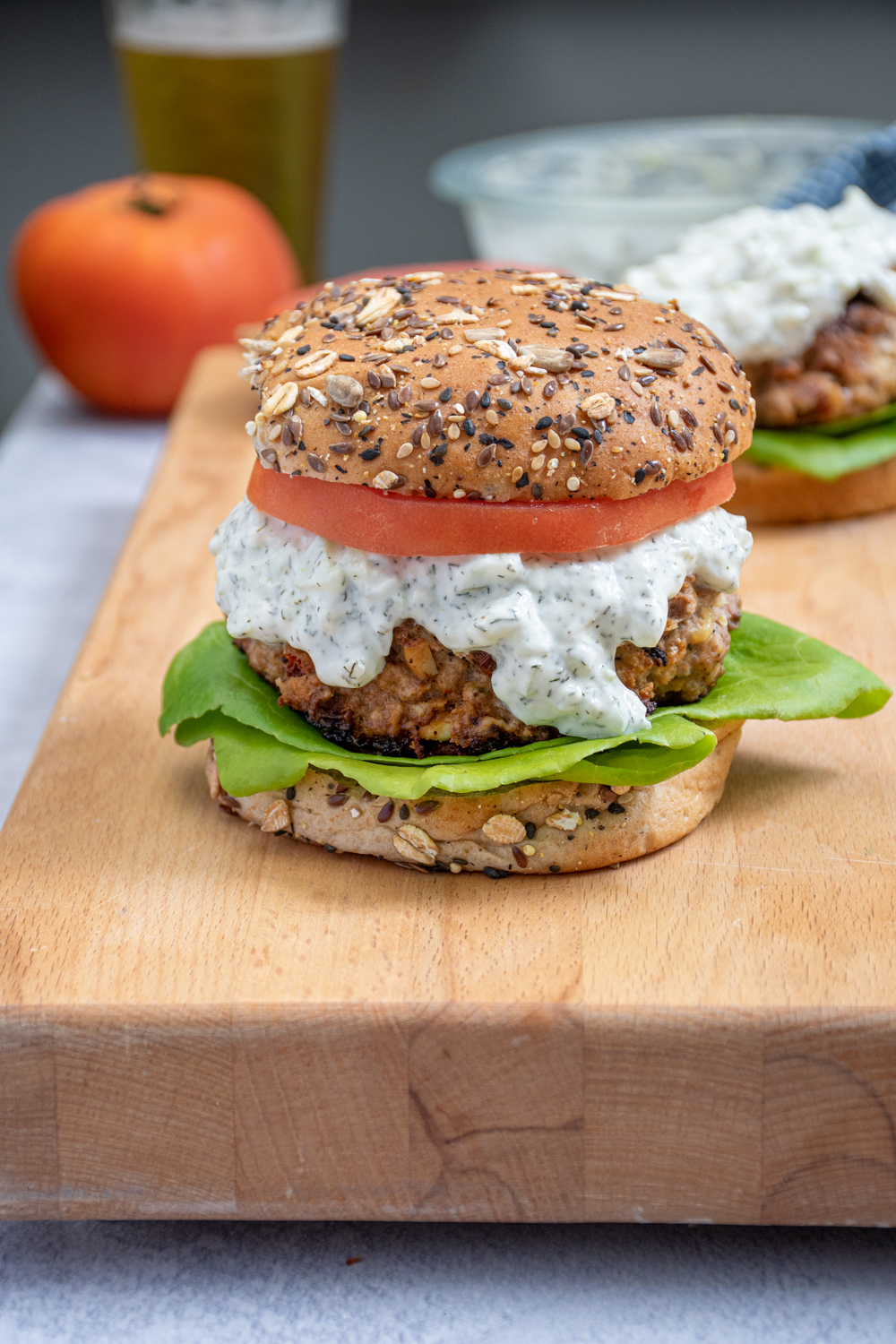 A Mediterranean Turkey Burger topped with Tzatziki sauce, tomatoes, and lettuce sitting on a butcher block next to another turkey burger.