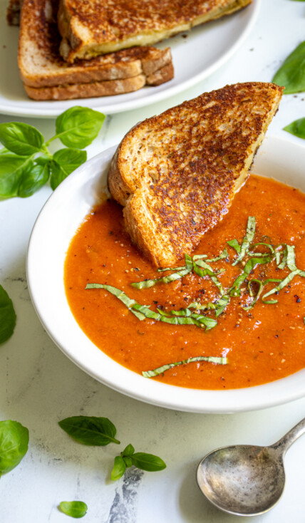 A white bowl filled with roasted red pepper and tomato soup garnished with fresh basil and a toasted grilled cheese being dunked into the soup.