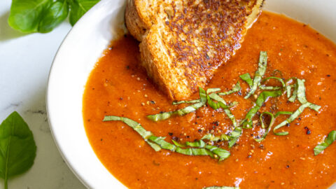 A white bowl filled with roasted red pepper and tomato soup garnished with fresh basil and a toasted grilled cheese being dunked into the soup.