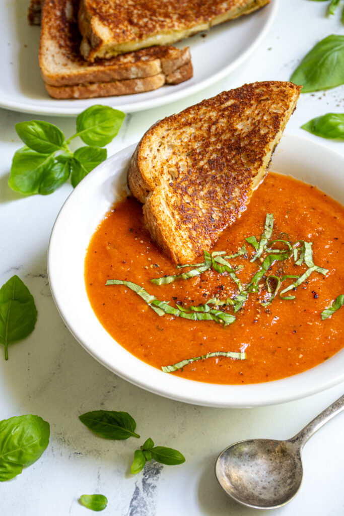 A white bowl filled with roasted red pepper and tomato soup garnished with fresh basil and a toasted grilled cheese being dunked into the soup.