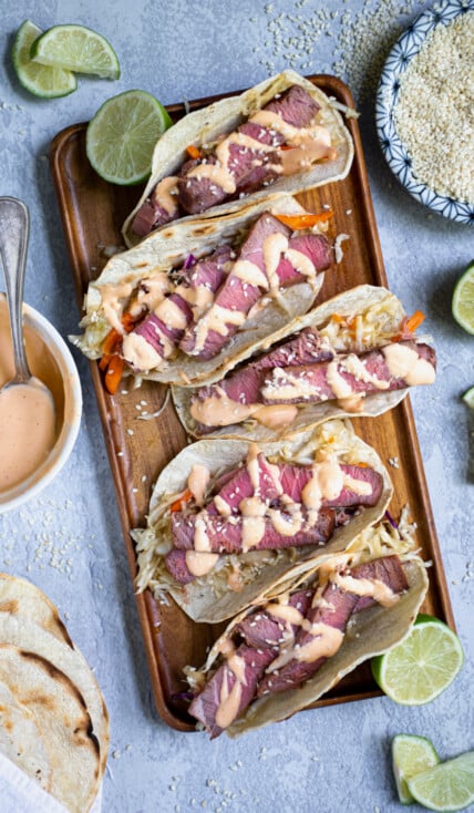 Five tuna steak tacos sitting on a string platter garnished with sriracha mayo and sesame seeds.