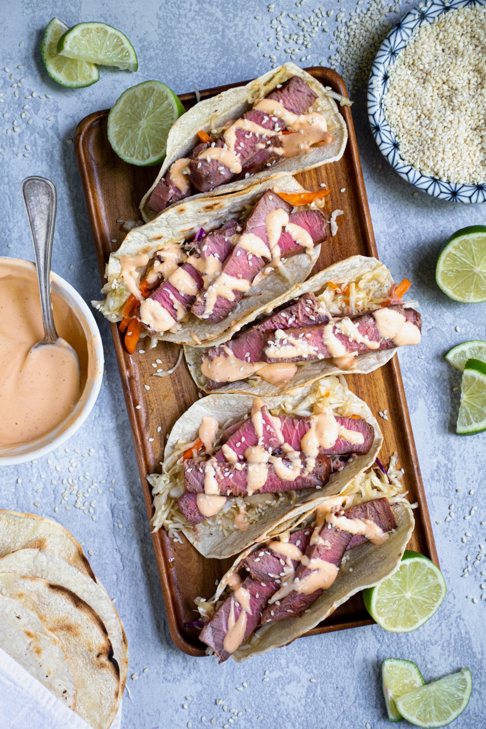Five tuna steak tacos sitting on a string platter garnished with sriracha mayo and sesame seeds.